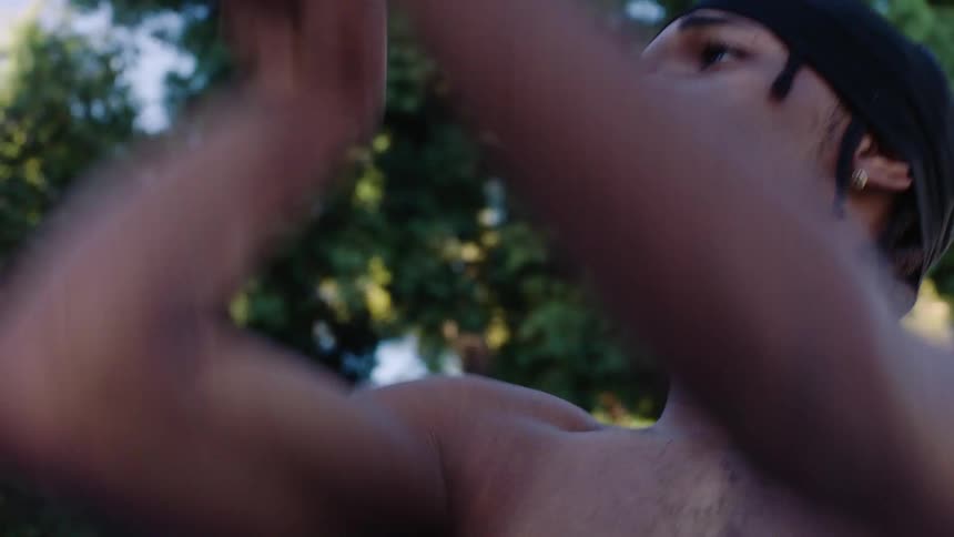 A shirtless man wearing a black durag looks toward a basketball hoop with a focused expression on an outdoor court surrounded by lush green trees