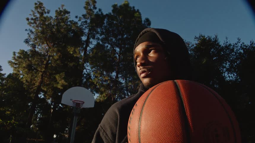 A young man in a black hoodie and durag holds a basketball close to his chest on an outdoor court he gazes off into the distance with a contemplative expression as the late afternoon sun highlights his face