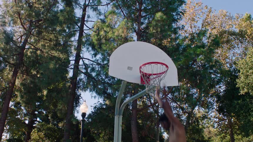 A shirtless man jumps and performs a slam dunk on an outdoor basketball court surrounded by tall green trees during a bright sunny day