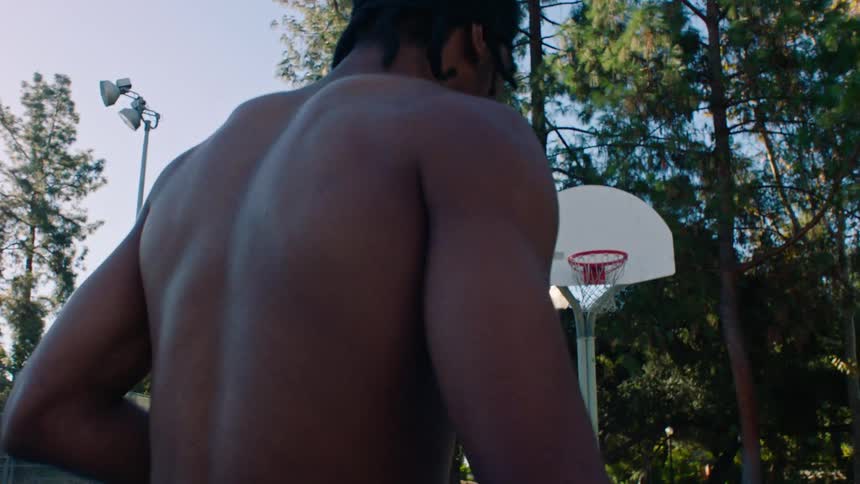 A young black athletic man playing basketball shirtless on an outdoor court at midday under bright sunlight