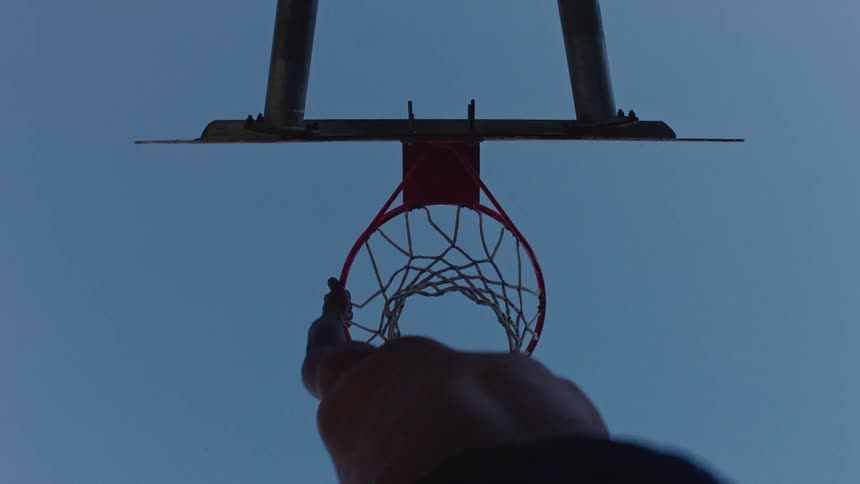 An athlete dunking a basketball into the hoop hanging from the rim for a few seconds before dropping down as the sunset casts a warm glow across the court