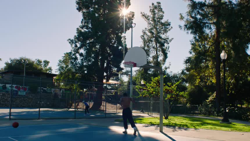 Person performs slam dunk on outdoor basketball court with sun shining through the trees
