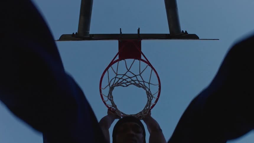 A man jumping to dunk a basketball briefly hanging on the hoop as the ball drops through all set against a sunset sky