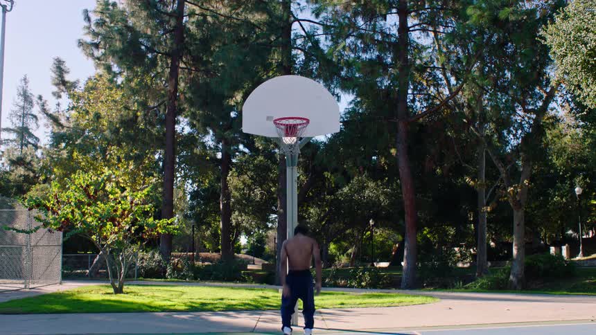 A shirtless man performs a powerful slam dunk on an outdoor basketball court surrounded by tall green trees and a clear blue sky during a sunny afternoon