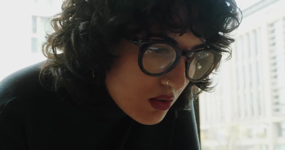 Young adult woman putting glasses on next to a window showing high rise buildings and an urban area outdoors during day time