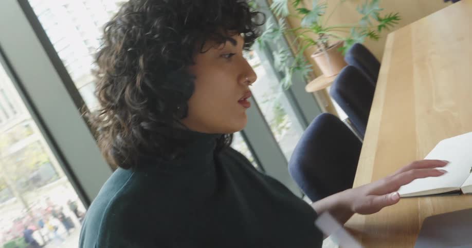 A woman with curly hair sits at a desk using a laptop while examining a small white wind turbine model in an office with large windows overlooking a city street