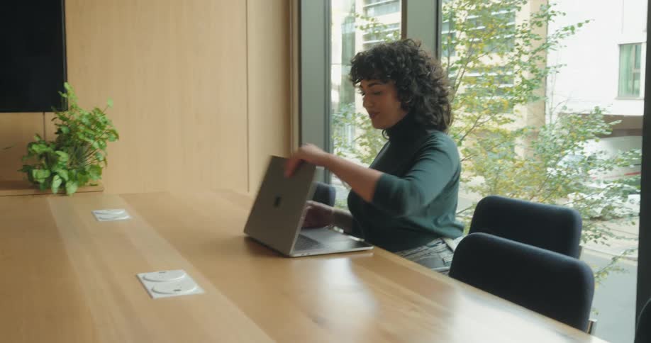 A woman with curly hair wearing a dark green turtleneck and jeans enters a modern office sits at a long wooden table by a large window and begins working on a silver laptop