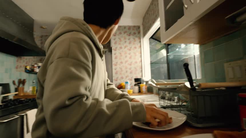 A young man in a grey hoodie quickly finishes eating puts on a black backpack and waves a brief goodbye before hurrying out of a kitchen decorated with floral wallpaper and blue tiles