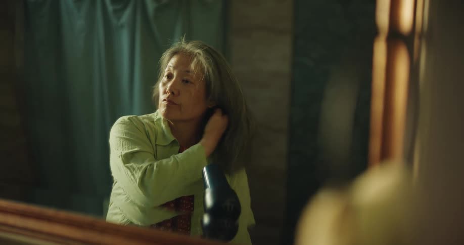 An older woman with grey hair stands before a bathroom mirror and uses a black dryer to style her hair while wearing a light green shirt over a patterned red top in soft indoor light