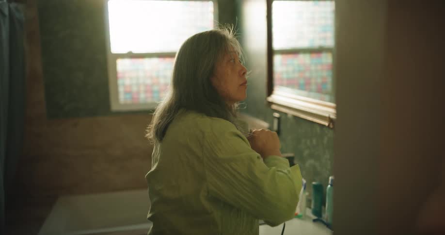 A mature woman stands in a sunlit bathroom looking into a mirror while using a black hairdryer to style her mediumlength gray hair surrounded by everyday toiletries and a decorative stained glass window