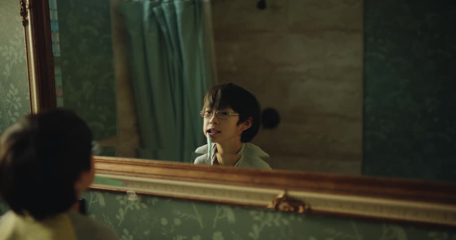 A young boy with glasses looks at his reflection in a bathroom mirror with an ornate gold frame before ducking out of view in a room with green floral wallpaper