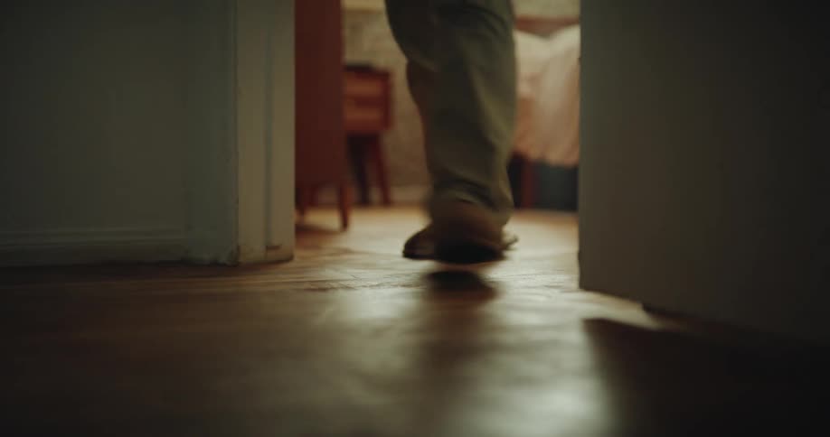 Person walks on wooden floor in domestic setting with soft lighting and figure in background
