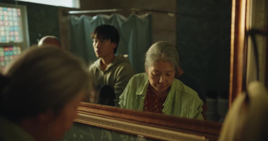 An elderly woman puts on her glasses while standing with three male family members of various ages as they look into a bathroom mirror and perform their morning routines together in a dimly lit room with floral wallpaper and a blue shower curtain