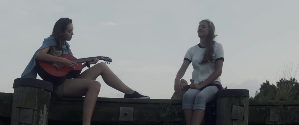 Two women sit on a wall outside, one playing guitar and one throwing something out of a paper bag