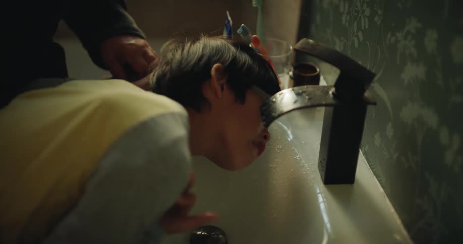 A young boy leans over a bathroom sink to rinse his mouth with water after brushing his teeth an older man with glasses stands beside him watching over the routine with a gentle smile