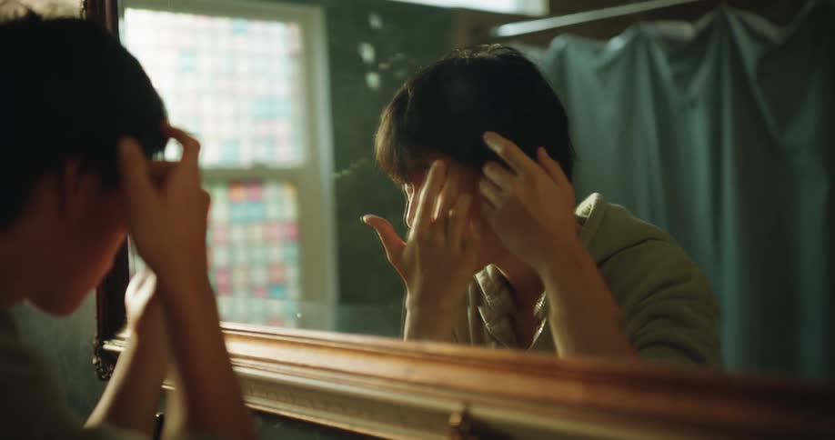 A young asian teenager wearing a green hoodie stands in front of a woodenframed mirror in a bathroom carefully adjusting his hair and inspecting his face while looking at his reflection during his morning routine