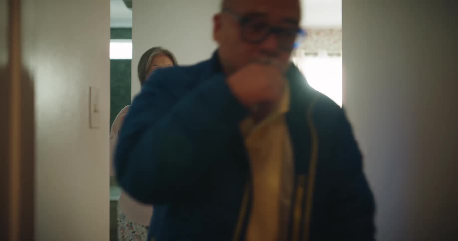 A senior man carrying a bag walks through a hallway and exits while a senior woman holding a mug waves goodbye to him from the doorway of a bedroom in a brightly lit home