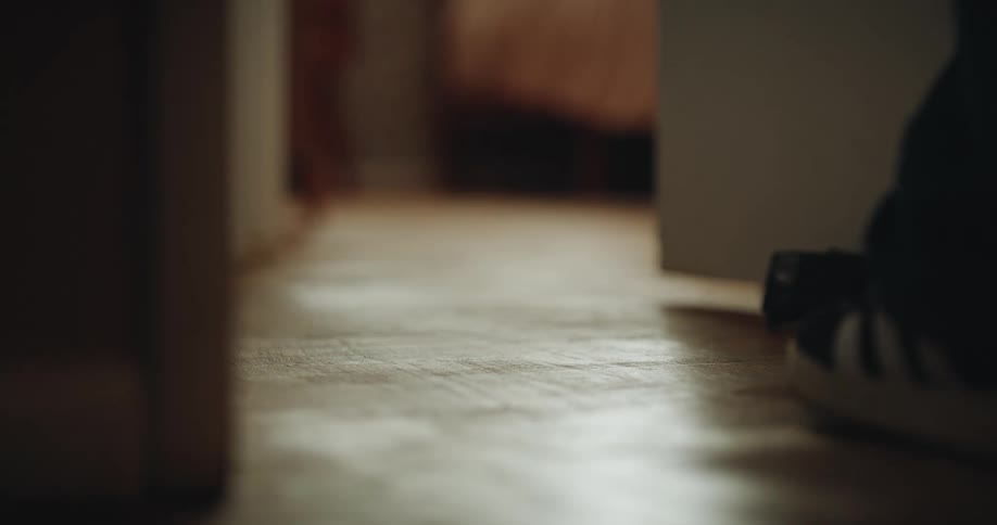 A low view of a child wearing black sneakers and dark trousers walking across a wooden floor through a doorway in a dimly lit room