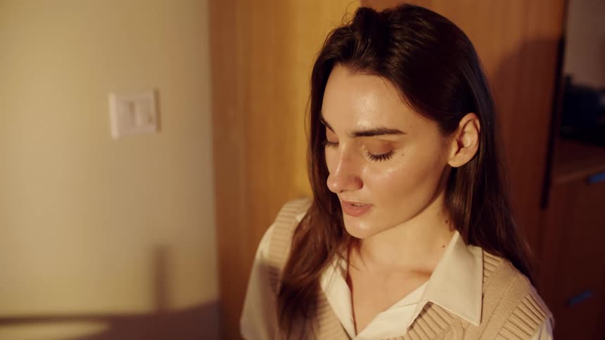 Person with long brown hair wears beige vest over white shirt in warmly lit indoor setting