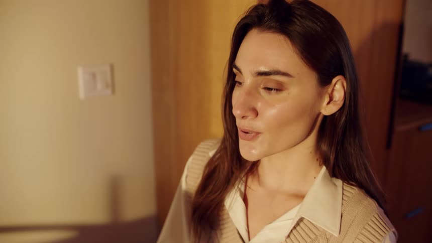 A woman with long brown hair wearing a beige sweater vest and white shirt smiles while looking in a dimly lit room with warm wood tones