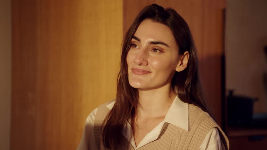 Woman indoors under warm lighting looks to the side with a neutral expression