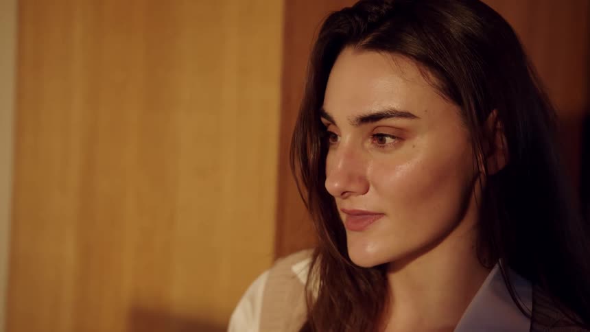 A closeup shot shows a young woman with long brown hair talking and changing her facial expressions while standing against a wooden wall in a room with warm lighting