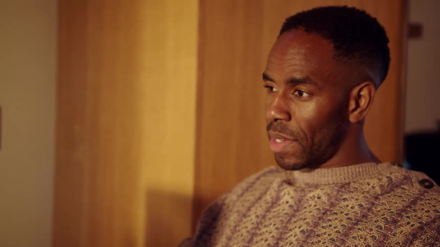 Young adult black man sitting and talking in front of a laptop computer over a wodden desk indoors during dusk or dawn