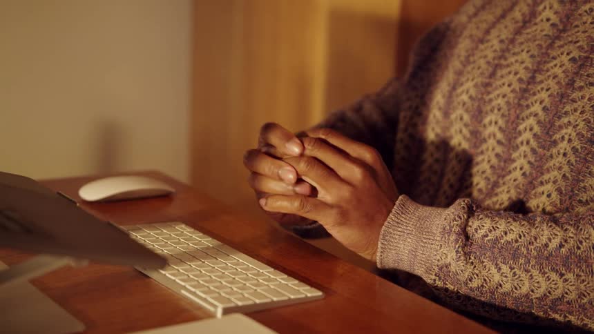 Interlocked hands rest on a wooden desk in front of a keyboard and laptop