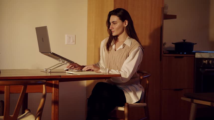 A woman wearing a tan vest over a white shirt sits at a wooden table and opens a laptop on a silver stand to begin working in a dimly lit room with a kitchen in the background
