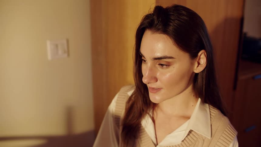 A woman with long brown hair wearing a beige sweater vest and white shirt smiles while looking in room with warm evening lighting