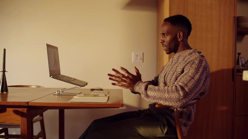 Man attends a video conference while seated at a table with a laptop placed on a stand