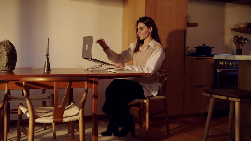 Woman seated at dining table works on laptop in cozy kitchen with decorative items and utensils in background