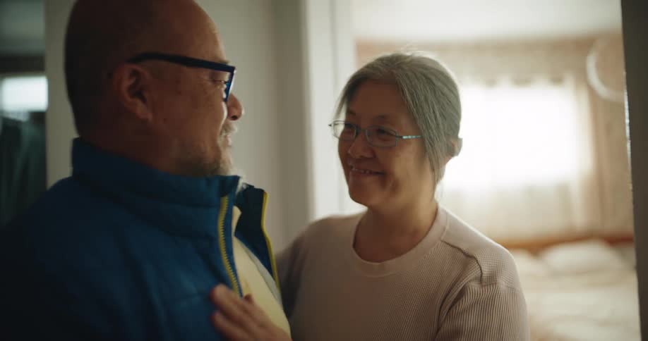 An elderly woman with gray hair and glasses gently helps an older man adjust his jacket and kisses him goodbye in a softly lit bedroom doorway