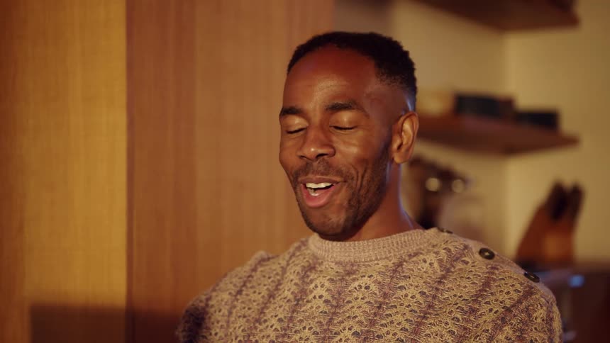 Man waves a hand while smiling in an indoor setting with warm lighting