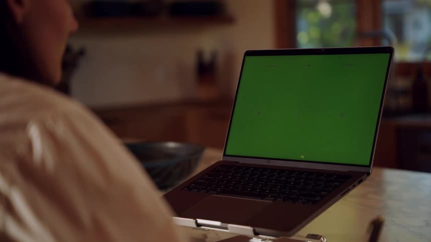 Woman sits at a table in front of a laptop computer with a green screen writing on paper