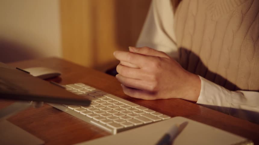 A woman sits at a wooden desk with her hands clasped before looking up and speaking toward a laptop screen in a room with warm lighting