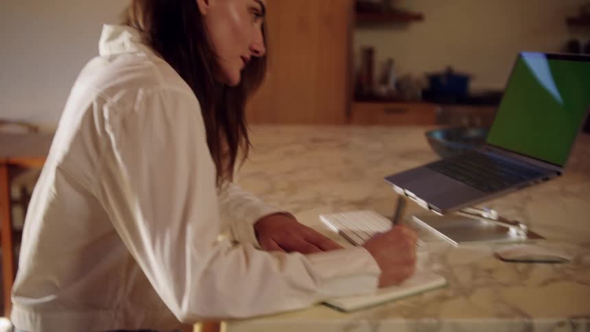 A young woman sitting in a dining area smiling as she writes on a notepad with her laptop open in front of her