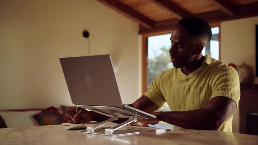 A man in a yellow shirt is sitting at a table focused on his laptop which is propped up on a silver stand as he works he alternates between typing on the keyboard and writing notes in a physical notebook beside him
