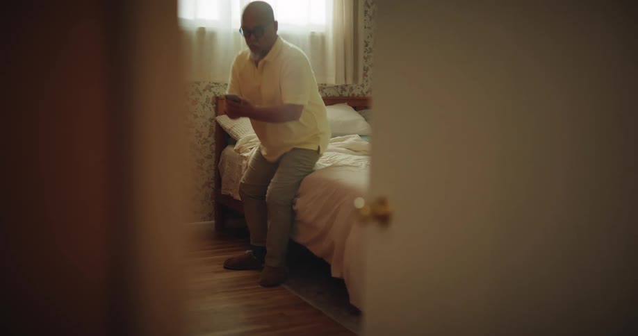 Adult man sitting down on a bed putting on socks on his feet then checking a smartphone and walking away carrying a backpack indoors during day time