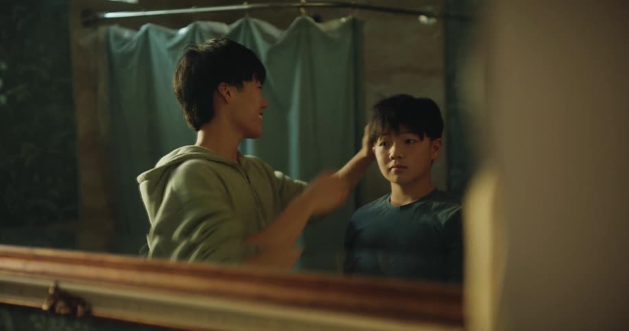 In a softly lit bathroom reflected through a mirror an older boy gently fixes a younger boys messy hair