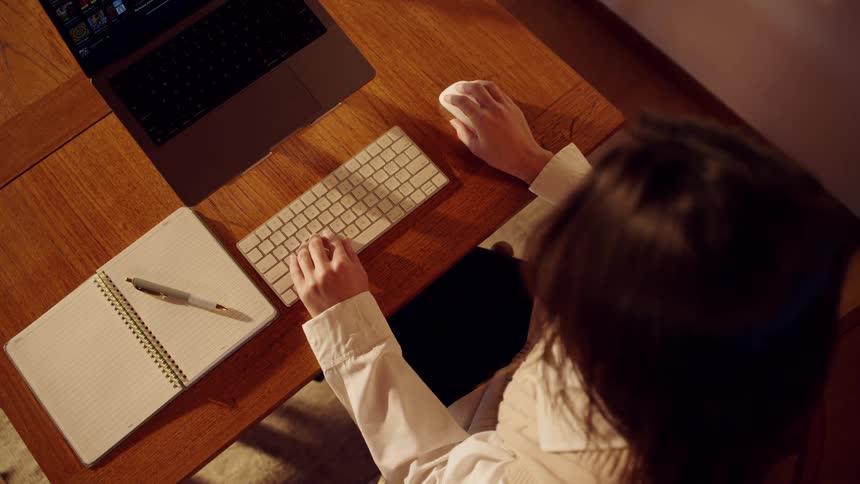 A person in a white shirt works at a wooden desk one hand on a mouse and the other typing on a keyboard beside a laptop with a neatly arranged notebook and pen nearby