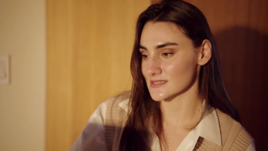 A woman wearing a sweater vest over a white shirt speaks and smiles while looking slightly offcamera toward a screen in a room with warm lighting and wooden wall panels