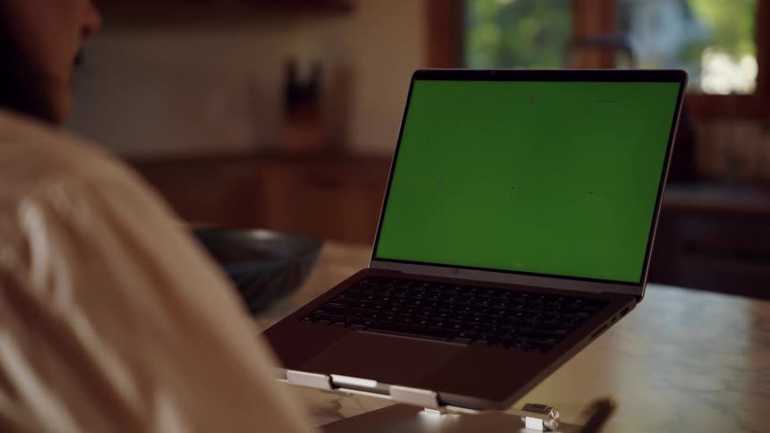 Woman sits at a table in front of a laptop computer with a green screen writing on paper