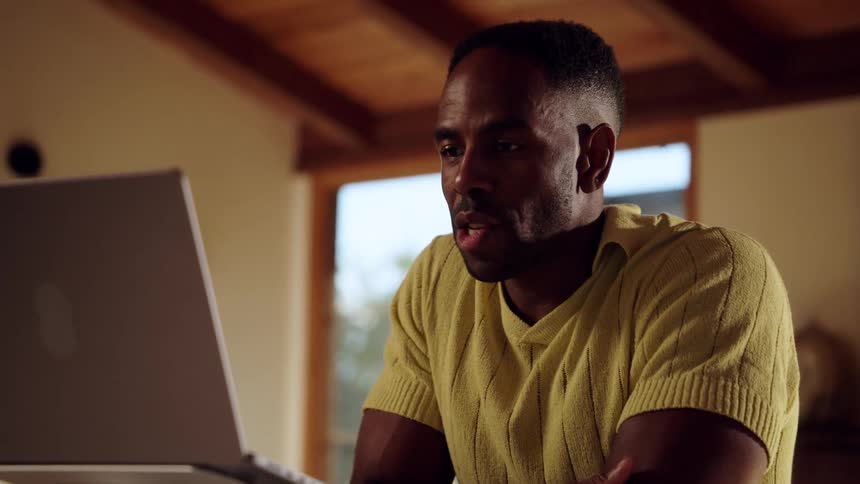 A man wearing a yellow knit shirt sits at a desk and writes in a notebook before looking at a laptop on a silver stand to engage in a video call inside a home with warm wooden ceilings