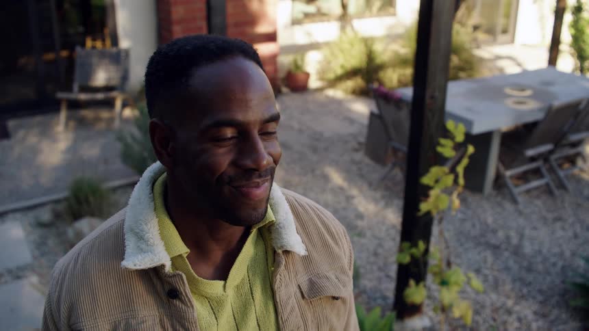 A man wearing a yellow shirt and a tan jacket with a fleece collar smiles while standing outdoors in a courtyard area he looks off to the side with a warm expression as sunlight illuminates the patio behind him