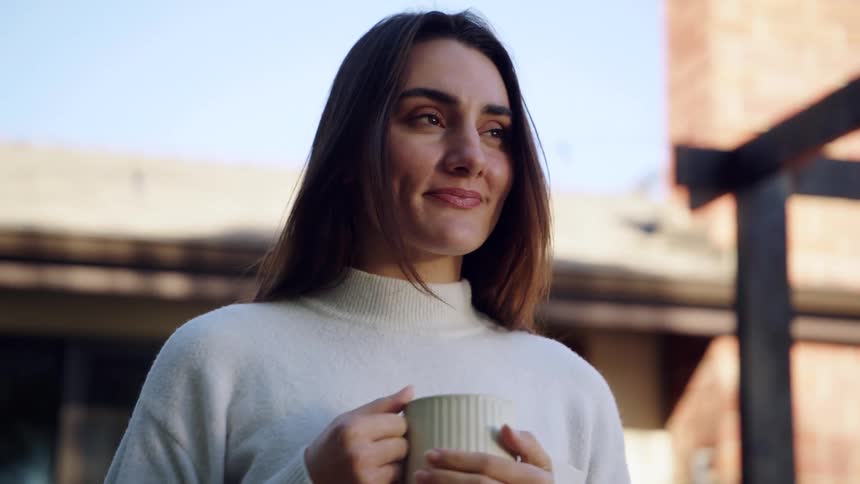 Woman in white sweater holds ceramic mug near face in sunlit outdoor setting