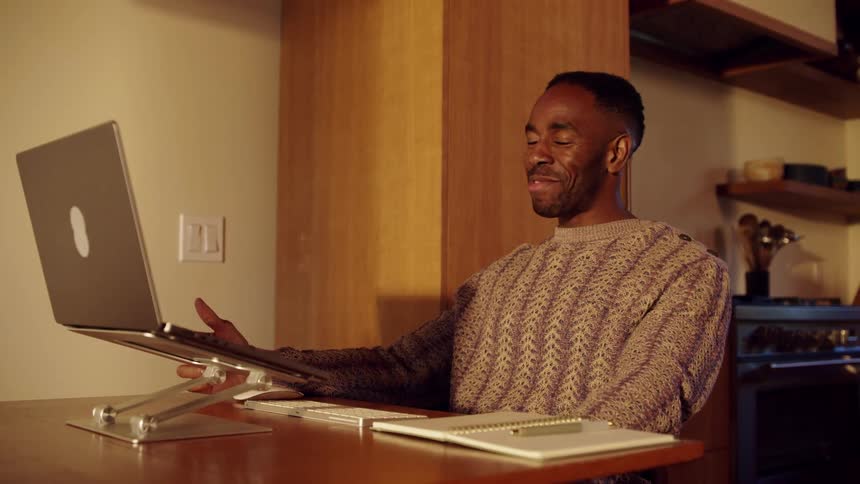 A black man talks to someone via his laptop while sitting in his dining room