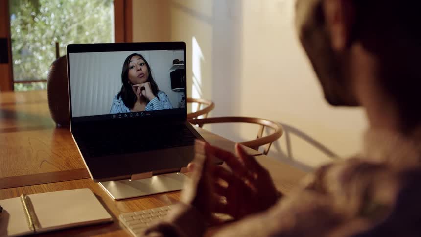 Young adult man talking in front of a laptop computer showing on the screen a young adult female indoors during dusk or dawn