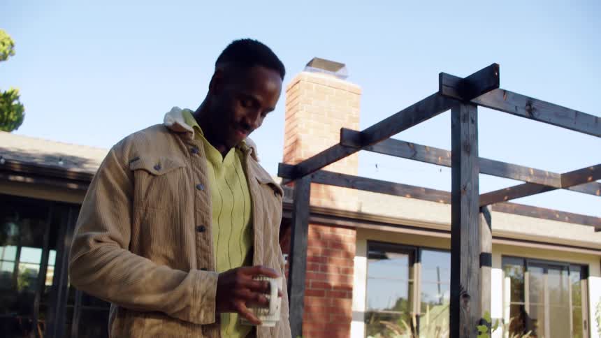 Man drinks a beverage from a cup while standing outdoors near a house