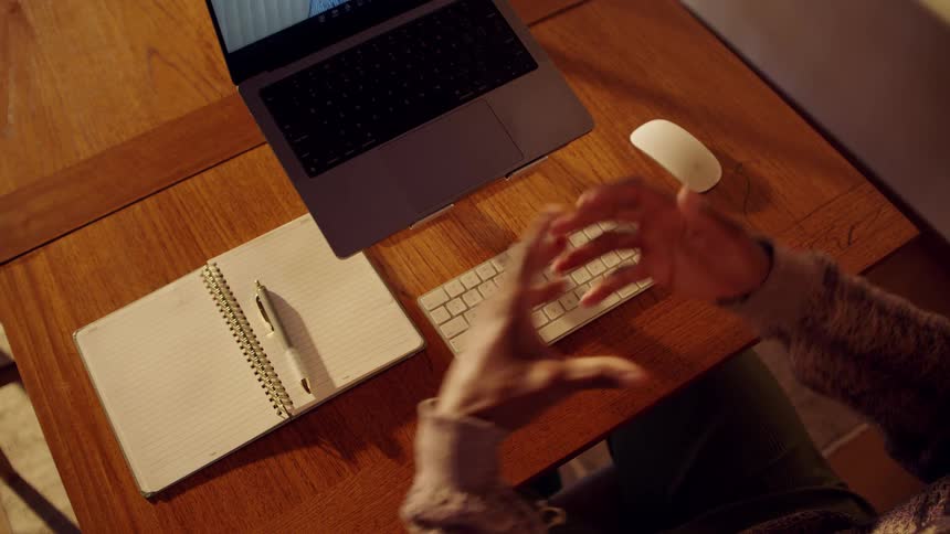 Person gestures while working on laptop at wooden desk in cozy indoor setting